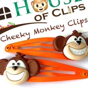 Puede incluir: Dos pinzas para el pelo de plástico naranja con caras de mono marrones. Los monos tienen ojos blancos, narices negras y dientes blancos. El texto "Cheeky Monkey Clips" está escrito sobre un fondo blanco.
