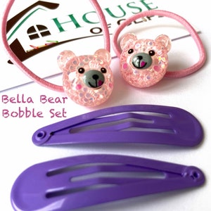 May include: A set of hair accessories featuring two pink bear bobbles with sparkly details and two purple plastic hair clips. The set is labeled "Bella Bear Bobble Set".