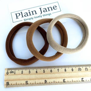 May include: Three hair scrunchies in brown and beige colors. The scrunchies are made of a soft, fuzzy material. The scrunchies are measured against a ruler, showing they are approximately 4 inches in diameter.