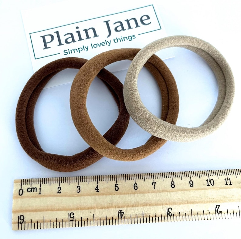 May include: Three hair scrunchies in brown and beige colors. The scrunchies are made of a soft, fuzzy material. The scrunchies are measured against a ruler, showing they are approximately 4 inches in diameter.