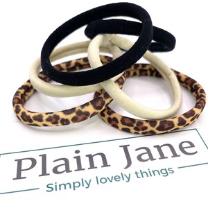 May include: A set of hair ties in a variety of colors and patterns, including black, beige, and leopard print. The hair ties are arranged in a pile on a white surface. The text "Plain Jane Simply lovely things" is visible below the hair ties.