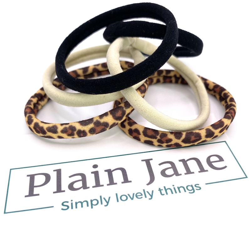 May include: A set of hair ties in a variety of colors and patterns, including black, beige, and leopard print. The hair ties are arranged in a pile on a white surface. The text "Plain Jane Simply lovely things" is visible below the hair ties.