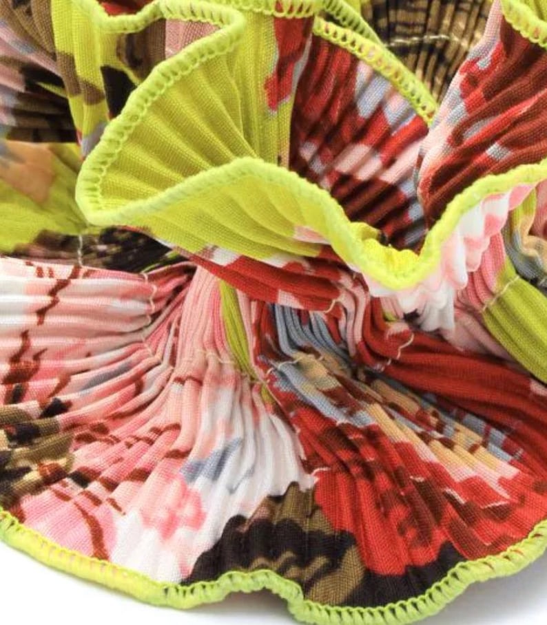 May include: Close-up of a pleated fabric with a floral print in shades of pink, red, white, and brown. The fabric has a lime green trim around the edges.