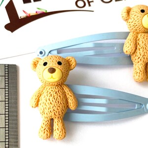 May include: Two light blue hair clips with brown teddy bear charms. The teddy bears have a yellow face with black eyes and a brown nose.