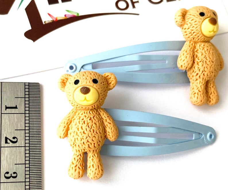 May include: Two light blue hair clips with brown teddy bear charms. The teddy bears have a yellow face with black eyes and a brown nose.