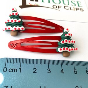 K&ouml;nnte beinhalten: Zwei rote Haarklammern mit einem gr&uuml;nen Weihnachtsbaum-Design. Jeder Baum hat wei&szlig;e und rote Punkte und einen braunen Stamm. Die Klammern werden mit einem Lineal gemessen.
