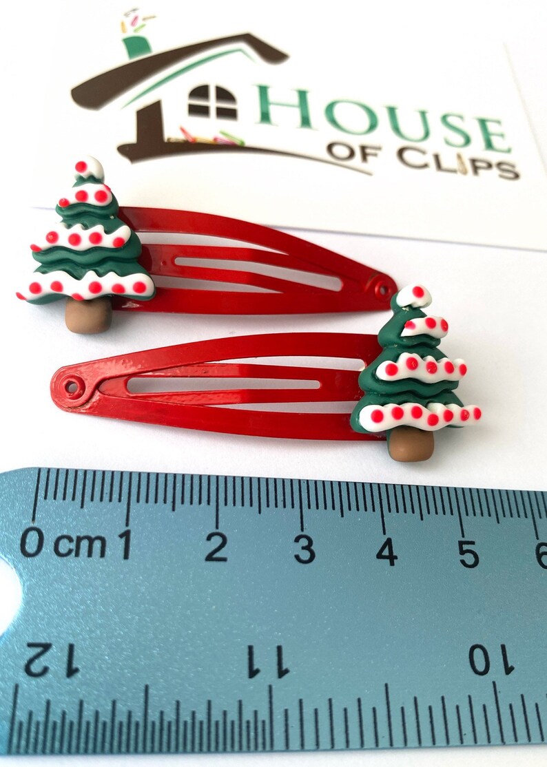 K&ouml;nnte beinhalten: Zwei rote Haarklammern mit einem gr&uuml;nen Weihnachtsbaum-Design. Jeder Baum hat wei&szlig;e und rote Punkte und einen braunen Stamm. Die Klammern werden mit einem Lineal gemessen.
