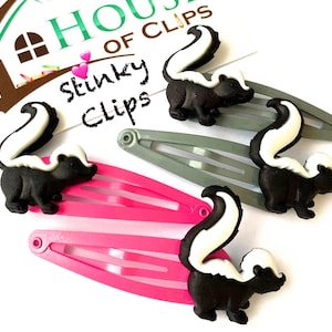 May include: Three black and white skunk hair clips. The clips are attached to pink and grey alligator clips. The clips have the text "Stinky Clips" on them.