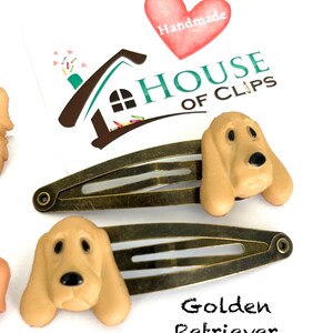 May include: Two bronze-toned metal hair clips, each adorned with a tan-colored Golden Retriever dog head. The clips are set against a white background, with the text "Golden Retriever" below. The image also includes a logo with the text "HOUSE OF CLIPS".