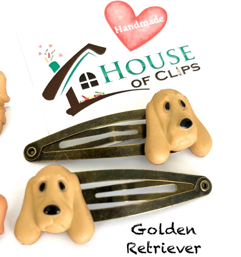 May include: Two bronze-toned metal hair clips, each adorned with a tan-colored Golden Retriever dog head. The clips are set against a white background, with the text "Golden Retriever" below. The image also includes a logo with the text "HOUSE OF CLIPS".
