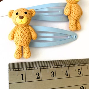 May include: Two light blue hair clips with brown teddy bear charms. The teddy bears are about 1 inch tall. The clips are measured against a ruler.