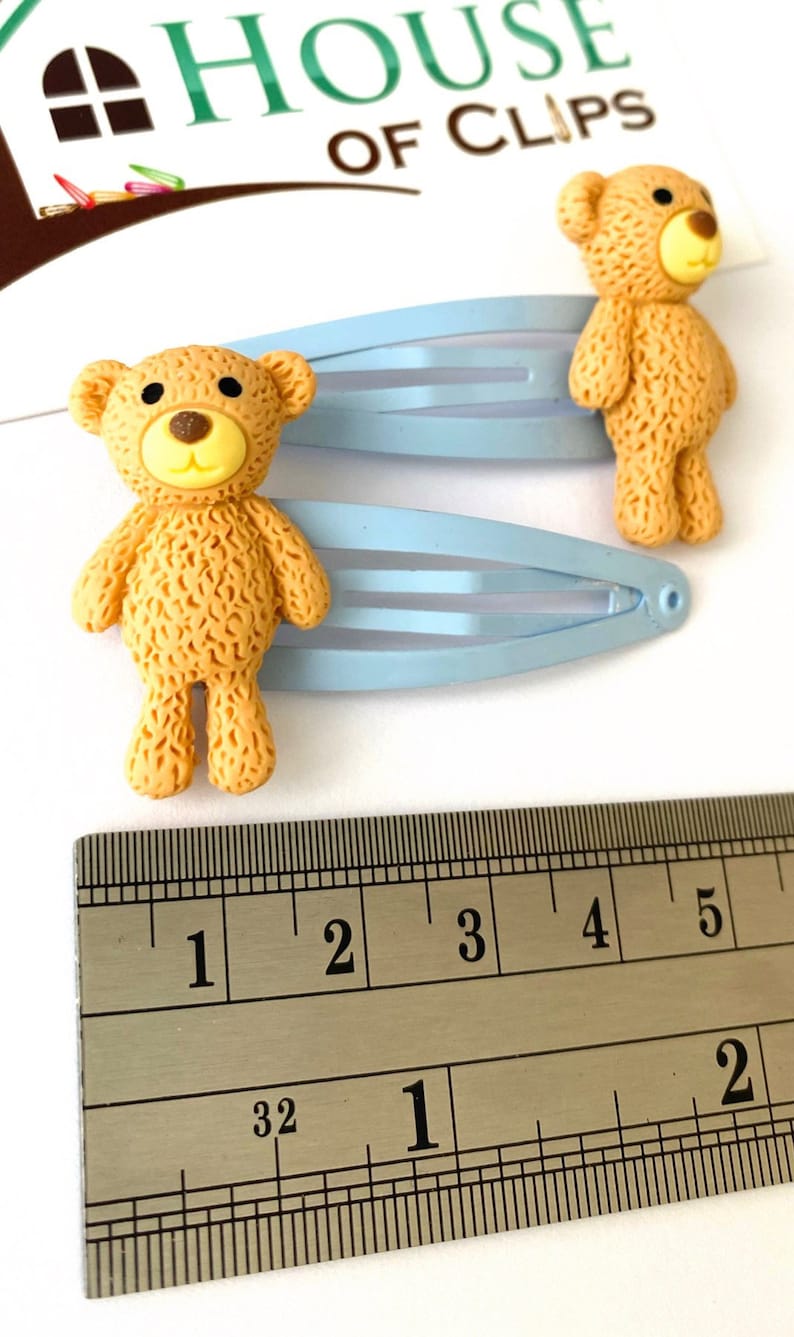 May include: Two light blue hair clips with brown teddy bear charms. The teddy bears are about 1 inch tall. The clips are measured against a ruler.