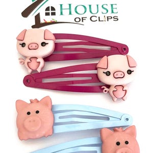 Könnte beinhalten: Set aus vier Haarklammern mit Schweinchen-Motiv. Zwei Klammern sind pink mit einem hellblauen Schweinchen-Design. Zwei Klammern sind hellblau mit einem pinken Schweinchen-Design. Die Klammern sind handgefertigt von House of Clips.