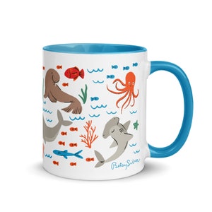 May include: White ceramic mug with a blue rim and handle. The mug features a colorful illustration of various sea creatures, including a walrus, octopus, hammerhead shark, and fish. The text "Betsy Silver" is printed on the bottom of the mug.