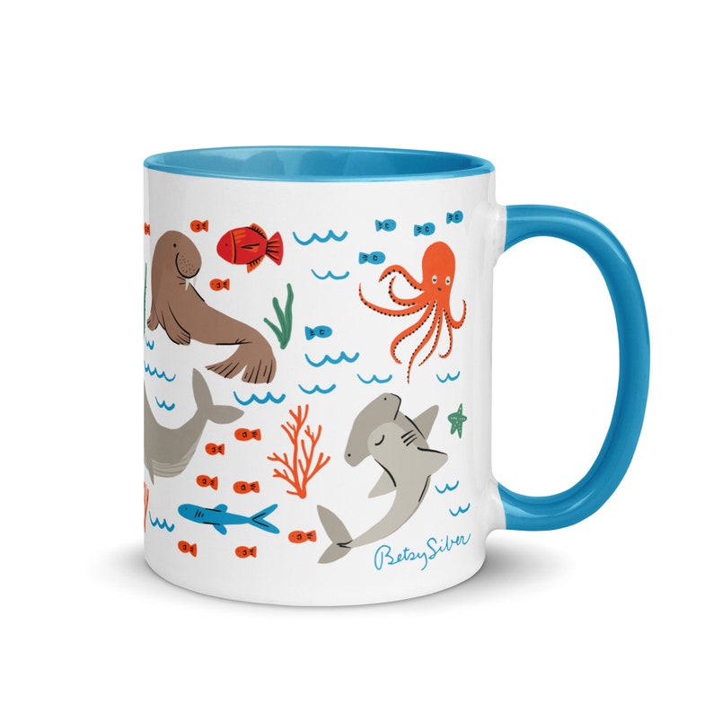 May include: White ceramic mug with a blue rim and handle. The mug features a colorful illustration of various sea creatures, including a walrus, octopus, hammerhead shark, and fish. The text "Betsy Silver" is printed on the bottom of the mug.