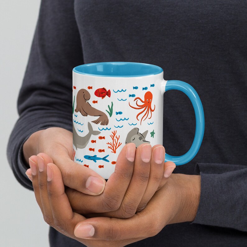 May include: A white ceramic mug with a blue rim and handle. The mug features a colorful illustration of various sea creatures, including a seal, an octopus, a shark, and fish. The background is white with blue waves.