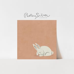 Könnte beinhalten: Illustration eines schlafenden weißen Kaninchens auf pfirsichfarbenem Hintergrund. Das Kunstwerk ist mit "Betsy Silver Illustration" in Schreibschrift signiert. Das Kaninchen ist zusammengerollt, die Augen geschlossen und die Ohren aufgestellt.