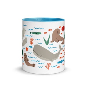 May include: A white ceramic mug with a light blue rim features a whimsical illustration of various sea creatures, including a whale, a walrus, and seals. The design also includes coral, seaweed, and fish.