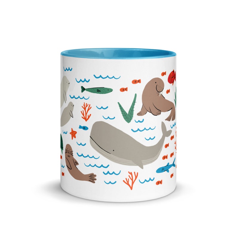 May include: A white ceramic mug with a light blue rim features a whimsical illustration of various sea creatures, including a whale, a walrus, and seals. The design also includes coral, seaweed, and fish.