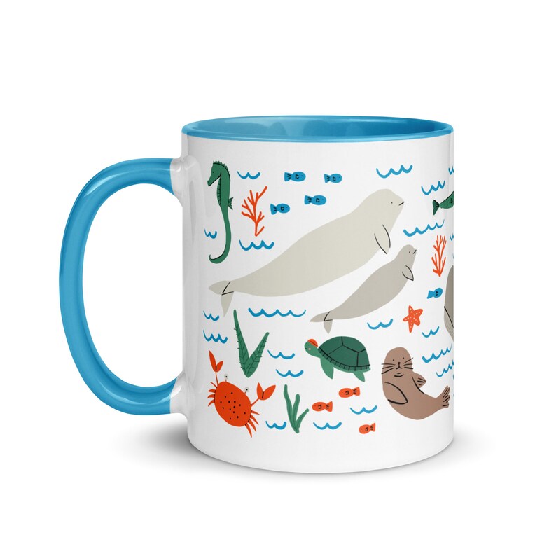 May include: White ceramic mug with a blue rim and handle. The mug features a colorful illustration of various sea creatures, including a seahorse, a crab, a turtle, a seal, and two walruses. The background is white with blue waves.