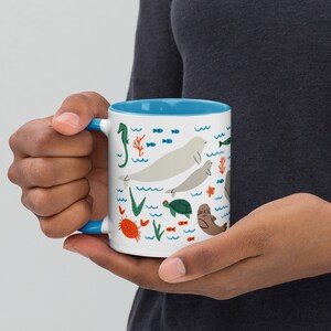 May include: A white ceramic mug with a blue rim features a colorful illustration of sea creatures, including a seahorse, fish, a turtle, a crab, and two walruses. The mug is held by a hand.