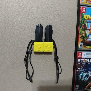 May include: A yellow question mark block wall mount with two black Nintendo Switch Joy-Con controllers attached. The mount has a black strap for hanging.