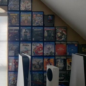 Può includere: Un muro con una grande collezione di custodie di videogiochi, disposte a forma di triangolo. Le custodie sono principalmente blu e nere, con alcuni accenti rossi e bianchi. Ci sono due console di gioco bianche su uno scaffale sotto il muro di custodie.