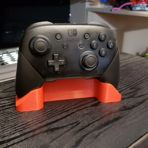 Op de afbeelding: Een zwarte Nintendo Switch Pro-controller op een rode plastic standaard. De controller heeft het Nintendo Switch-logo en de woorden "Nintendo Switch" erop gedrukt.