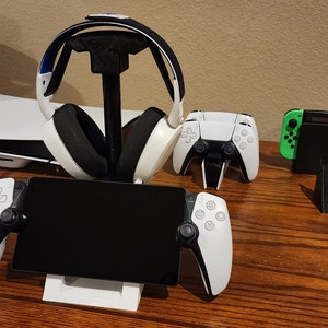 Puede incluir: Un auricular de juego blanco con una diadema negra está en un soporte. Hay dos mandos inalámbricos blancos y una consola Nintendo Switch negra sobre una mesa.