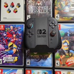 May include: A black Nintendo Switch controller is centered in front of a collection of video game cases. The controller has blue and red buttons and the Nintendo Switch logo. Game titles include "Doom", "Octopath Traveler", and "Ring Fit Adventure".
