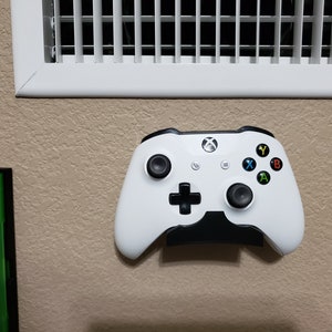 May include: A white Xbox controller is mounted on a wall with a black wall mount. The controller has black buttons and a black d-pad. The controller is in a vertical position.