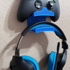 May include: A blue and black gaming headset with a microphone is hanging on a wall mount next to a black Xbox controller.