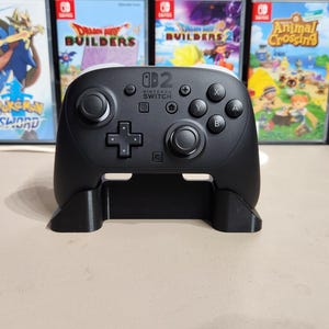 May include: A black Nintendo Switch Pro Controller with a matching stand. The controller has a D-pad, face buttons, and analog sticks. The Nintendo Switch logo is visible on the controller. Several Nintendo Switch game cases are in the background.