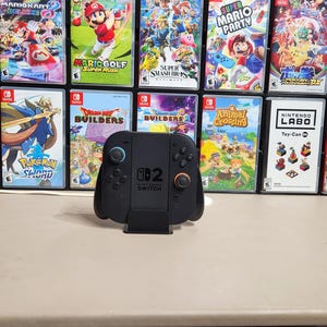 Op de afbeelding: Een zwarte Nintendo Switch-controller met blauwe en rode knoppen wordt getoond voor een verzameling game-cases. De cases hebben kleurrijke graphics en titels als "Mario Odyssey" en "Just Dance 2022."