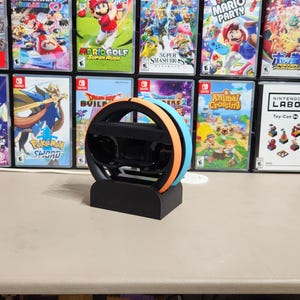 Può includere: Un volante da gioco nero con dettagli arancioni e blu, appoggiato su una base nera. È posizionato di fronte a una collezione di custodie di giochi per Nintendo Switch, tra cui i titoli "Just Dance" e "Mario Party".