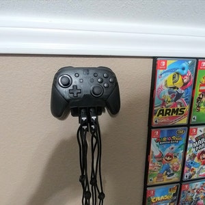 May include: A black video game controller mounted on a wall, with multiple cords extending downwards. The controller is positioned above a display of colorful video game cases, showcasing various titles.
