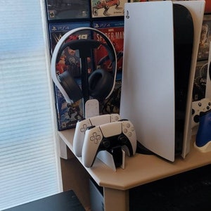 Puede incluir: Una consola PlayStation 5 blanca con un acento negro, dos mandos blancos y unos auriculares negros sobre un soporte. Se ven cajas de videojuegos en el fondo.