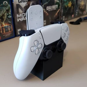 May include: A white PlayStation 5 DualSense controller is mounted on a black stand. A black remote control is also mounted on the stand.