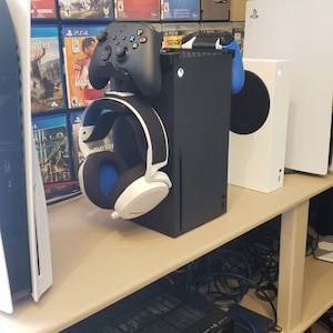 May include: A black Xbox Series X console with a white headset and a black Xbox controller on a shelf with a white PlayStation 5 console and a white PlayStation 4 console. The shelf is filled with video game cases.