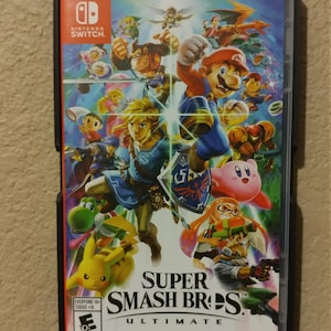 Puede incluir: Una caja de juego de Nintendo Switch con un borde negro y una imagen colorida de personajes del juego Super Smash Bros. Ultimate. El juego está clasificado como E para todos por la ESRB.