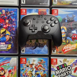 May include: A black Nintendo Switch controller rests on a display of various game cases. The cases feature colorful graphics and titles like "Marvel Ultimate Alliance 3" and "Ring Fit Adventure." The controller has a directional pad, buttons, and analog sticks.