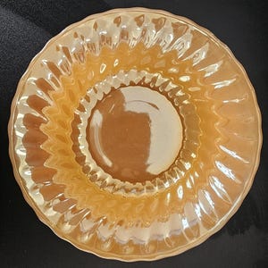 May include: A vintage, iridescent, amber-colored glass plate. The plate has a circular shape with a rippled, scalloped edge. The center of the plate is a solid amber color, and the edges have a textured, wavy design.