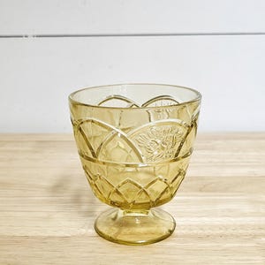 May include: A vintage amber-colored glass dessert dish with a pedestal base. The bowl features a diamond and floral pattern. The glass has a slightly textured appearance. The dish is empty and sits on a wooden surface.