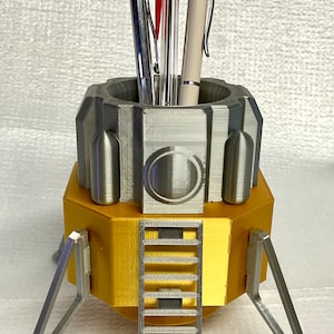 May include: A 3D printed silver and gold lunar lander pen holder with a ladder on the side and three legs. The pen holder has a silver top with a circular design and is holding three pens, one red, one silver, and one white.