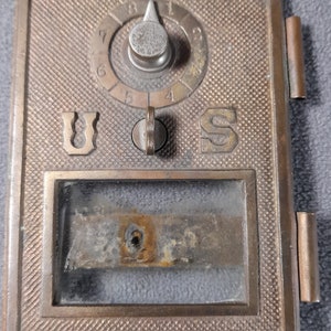 Puede incluir: Un buzón estadounidense de latón con una ventana de vidrio y un dial circular con los números del 1 al 8. El buzón tiene una inscripción de U.S.