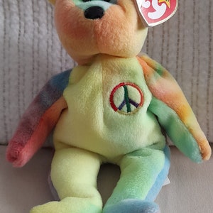 Könnte beinhalten: Ein Plüschtierbär im Batik-Look mit einem Peace-Zeichen auf der Brust. Der Bär sitzt auf einer weißen Oberfläche und hat ein rotes "Beanie Baby"-Etikett am Ohr.