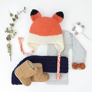 May include: A crocheted fox hat with orange and white stripes and black ears. The hat has long braided ties. The hat is laying on a gray shirt with a navy blue crocheted scarf and brown booties.