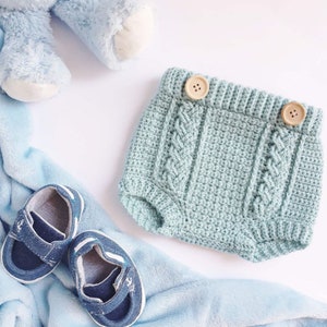Peut inclure: Une barboteuse bébé en crochet bleu clair avec des boutons en bois. La barboteuse a un motif en tricot torsadé et est posée sur une surface blanche.