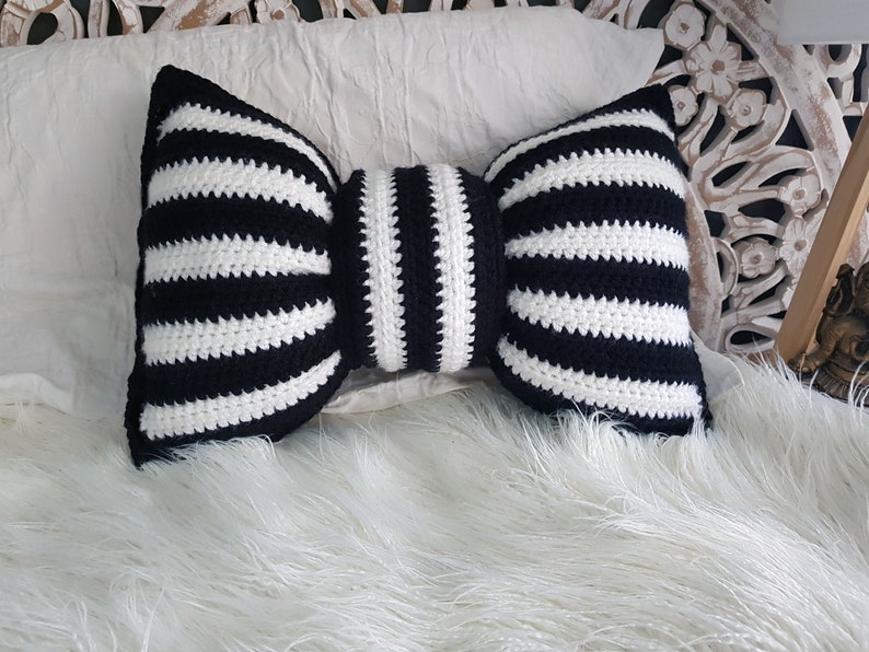 Pu&ograve; includere: Un cuscino a forma di fiocco in crochet a righe bianche e nere. Il cuscino &egrave; su una superficie bianca e soffice.