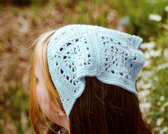 May include: A light blue crocheted bandana with a delicate openwork pattern. The bandana is tied around the head of a person with long brown hair.